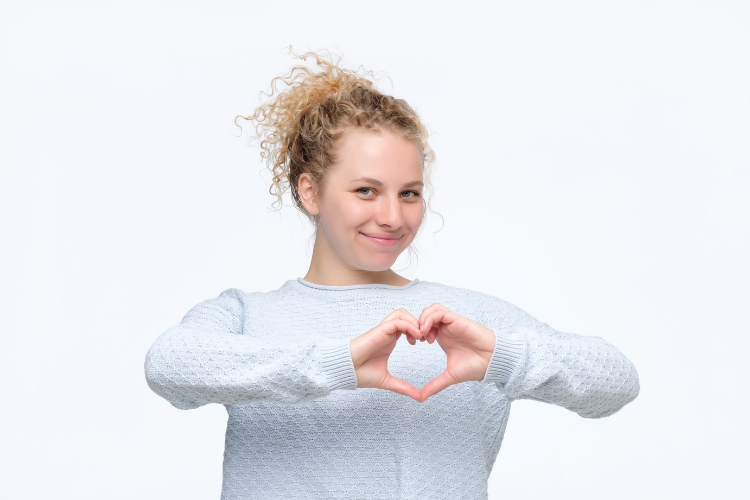 Jonge vrouw met opgestoken blond haar, witte trui met lange mouwen, maakt met haar handen het hartjesteken.